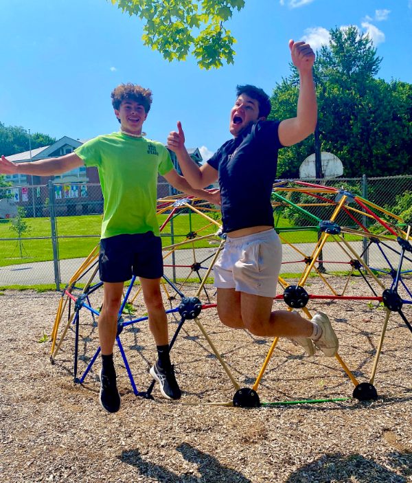 Two park coordinators jumping in mid-air outside in front of a multi-colored geometric climbing dome