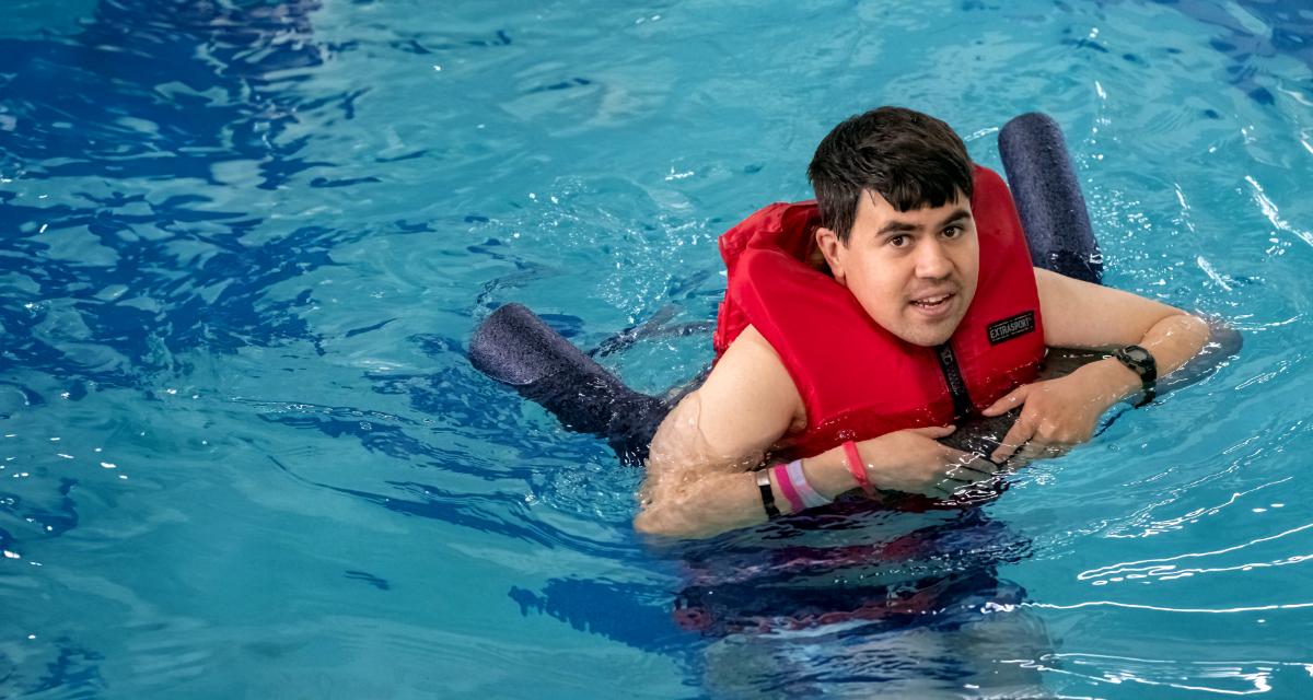 2024 Swim-A-Thon Ambassador Josh swimming in a pool with a red life vest and black pool noodle
